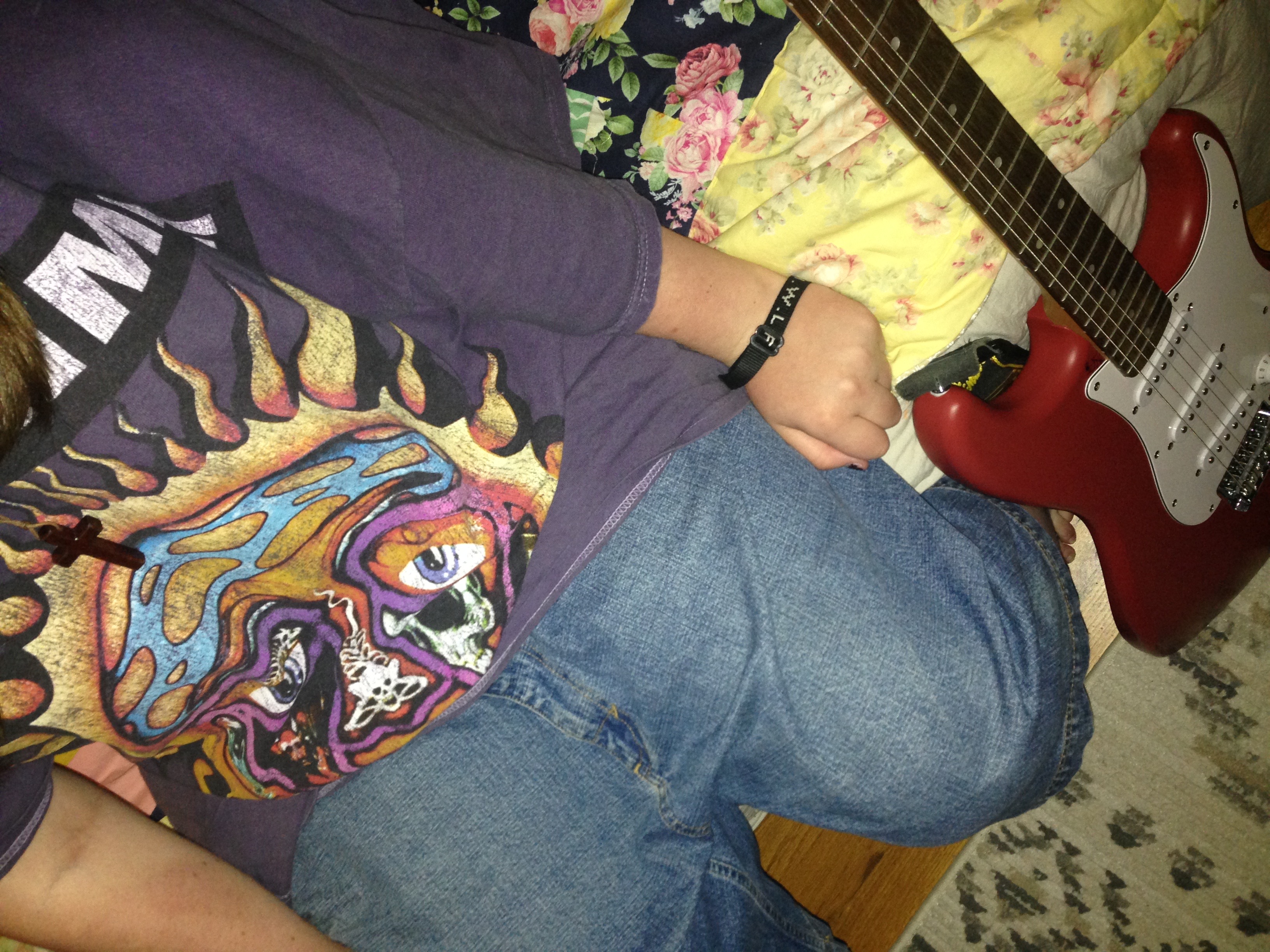 girl with brown hair wearing blue jorts, a sublime t-shirt, and a necklace with a cross on it sitting beside her guitar on her bed, and her sister is taking her photo.